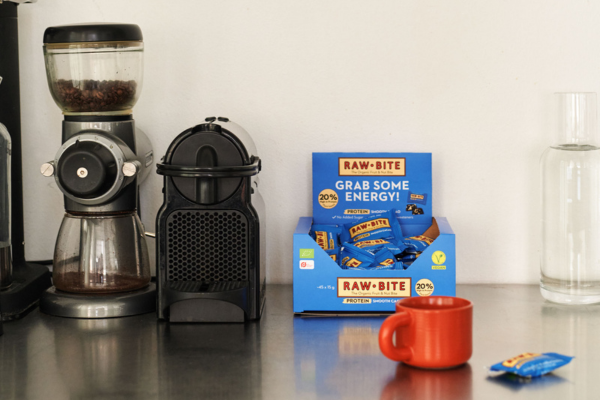 Coffee grinder, coffee press, and Rawbite Protein Smooth Cacao Office Box on a kitchen counter.