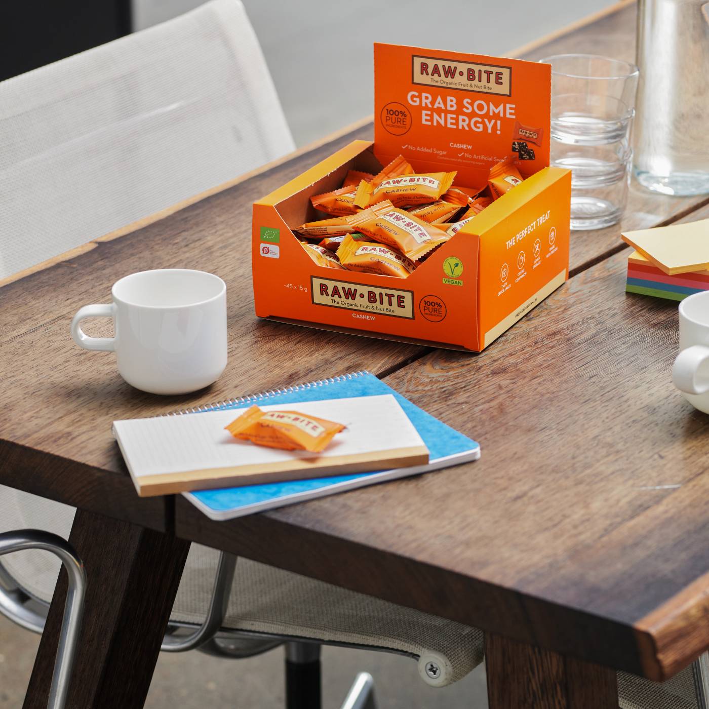 Box of RAWBITE mini cashew bars on a wooden table with a notebook and cups.