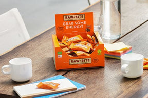 Box of RAWBITE cashew mini bars on a table with mugs and snacks.