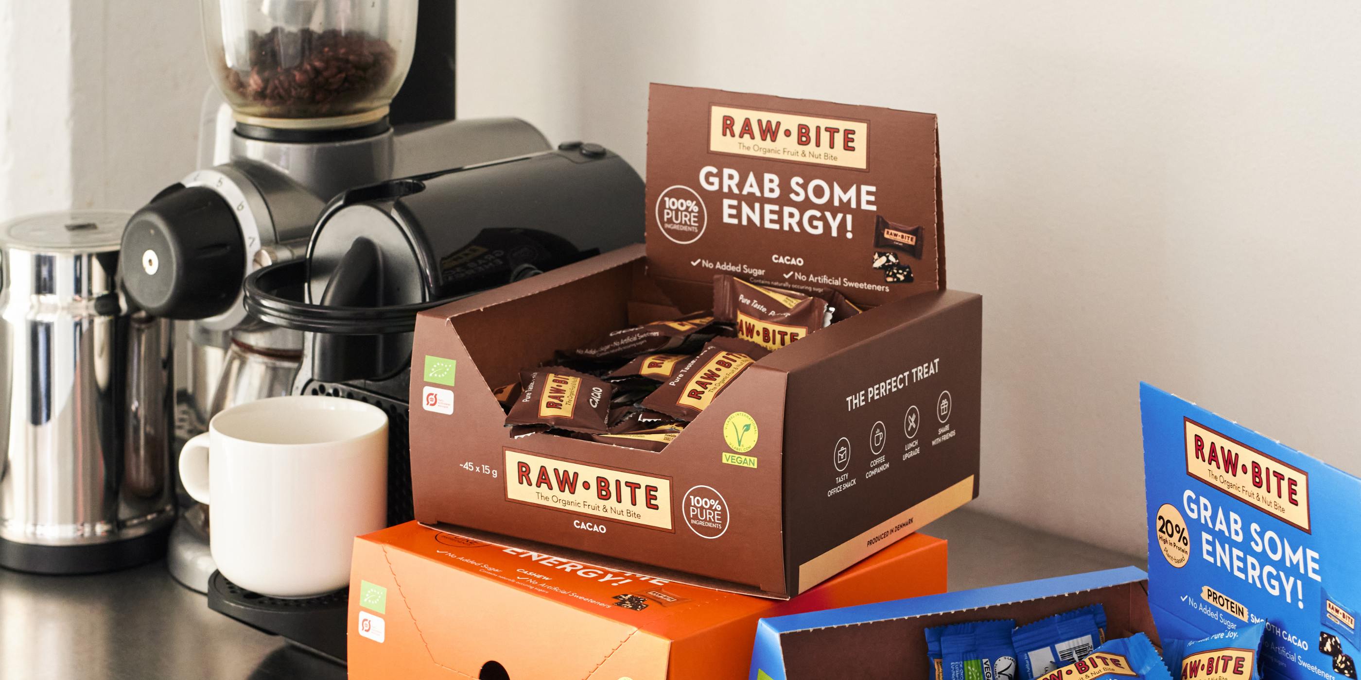 Rawbite mini bars displayed in front of a coffee machine with a white background