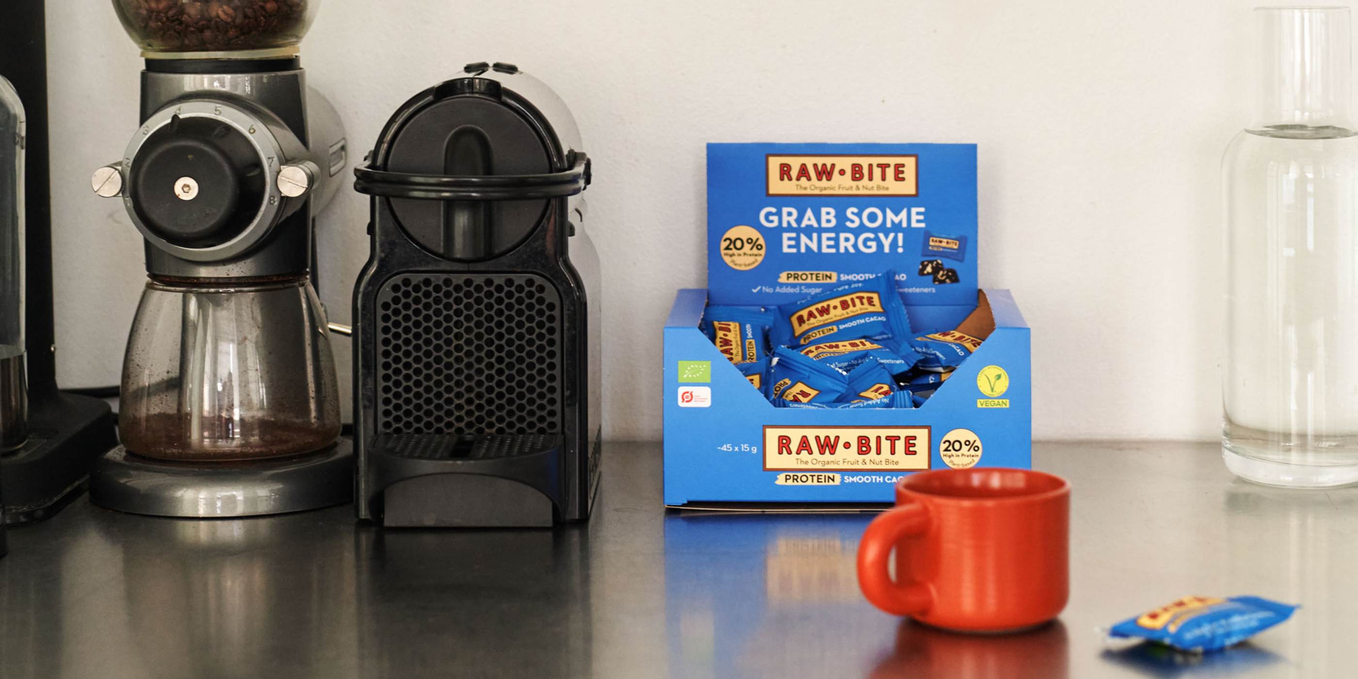 RAWBITE Protein Smooth Cacao Office Box on a kitchen counter with coffee-making equipment and a mug.