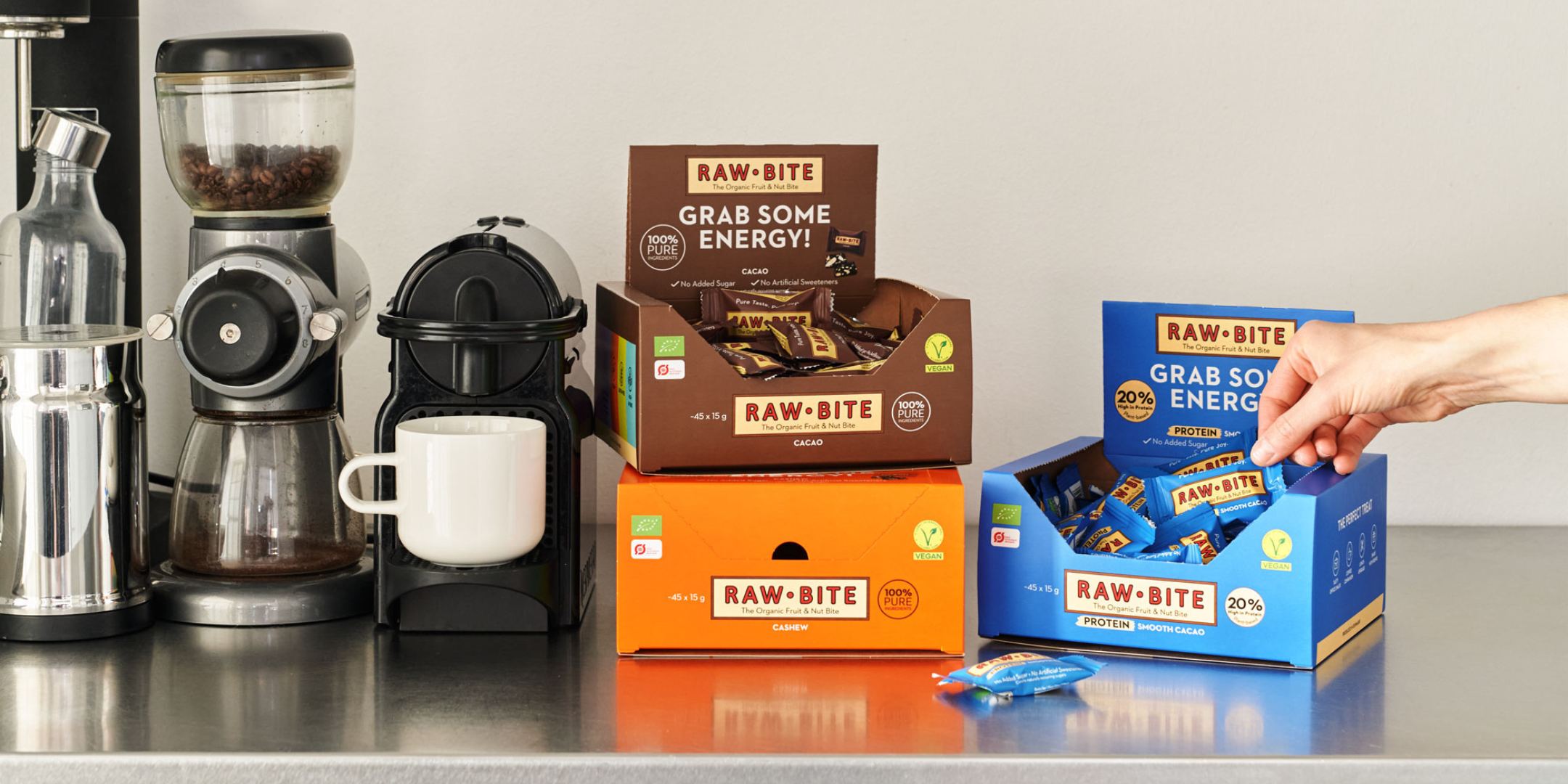 RAWBITE bars in various flavors on a kitchen counter with coffee-making equipment.