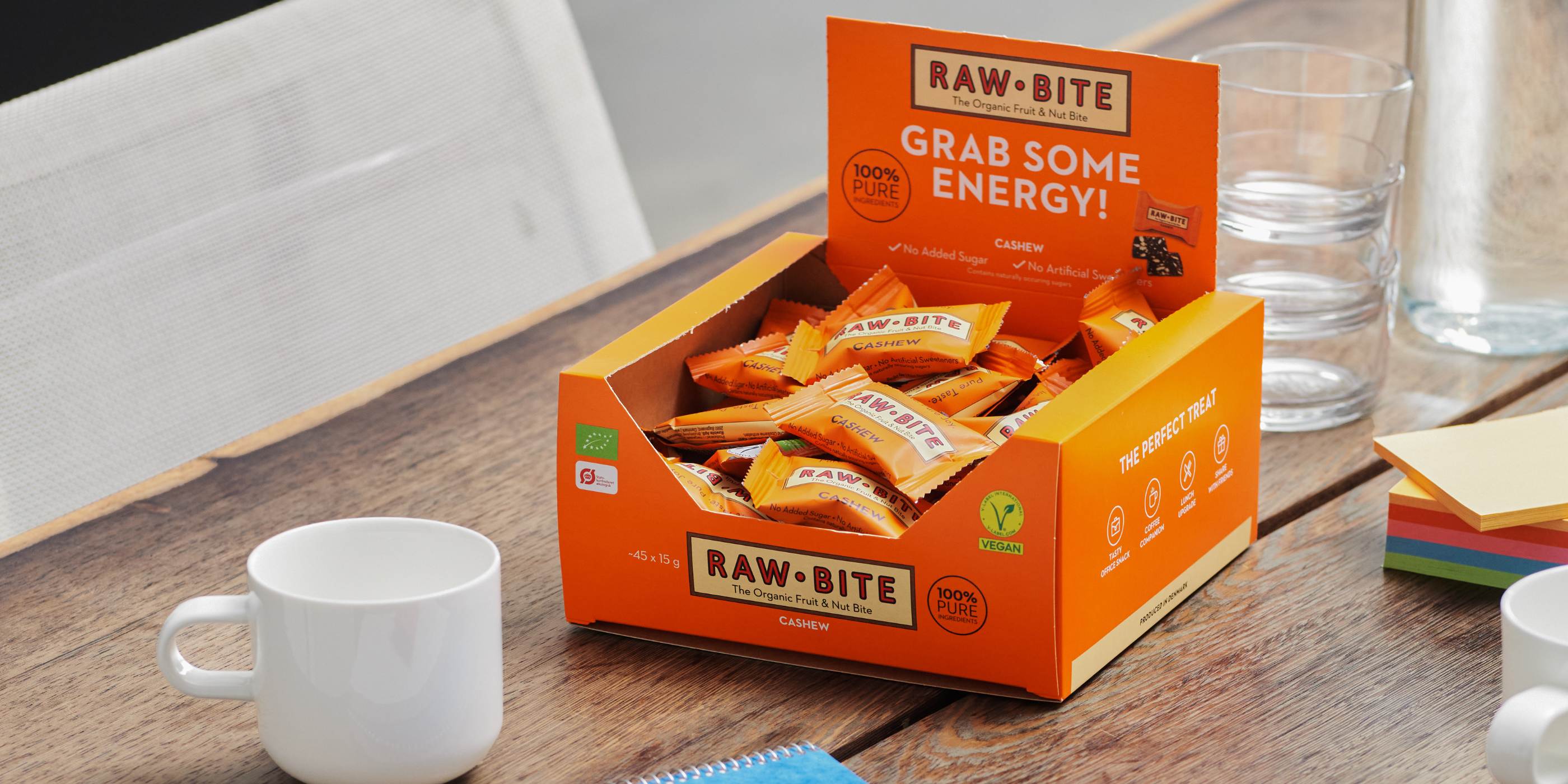 Open RAWBITE Office Box with Cashew mini bars on a table with a cup and water glasses.