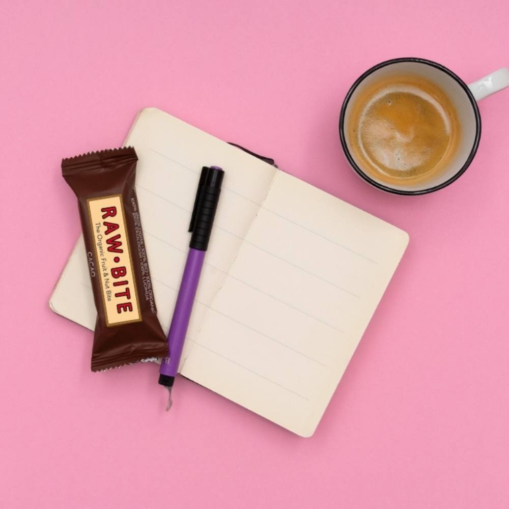 Open notebook with a RAWBITE bar, pen, and coffee cup on a pink background